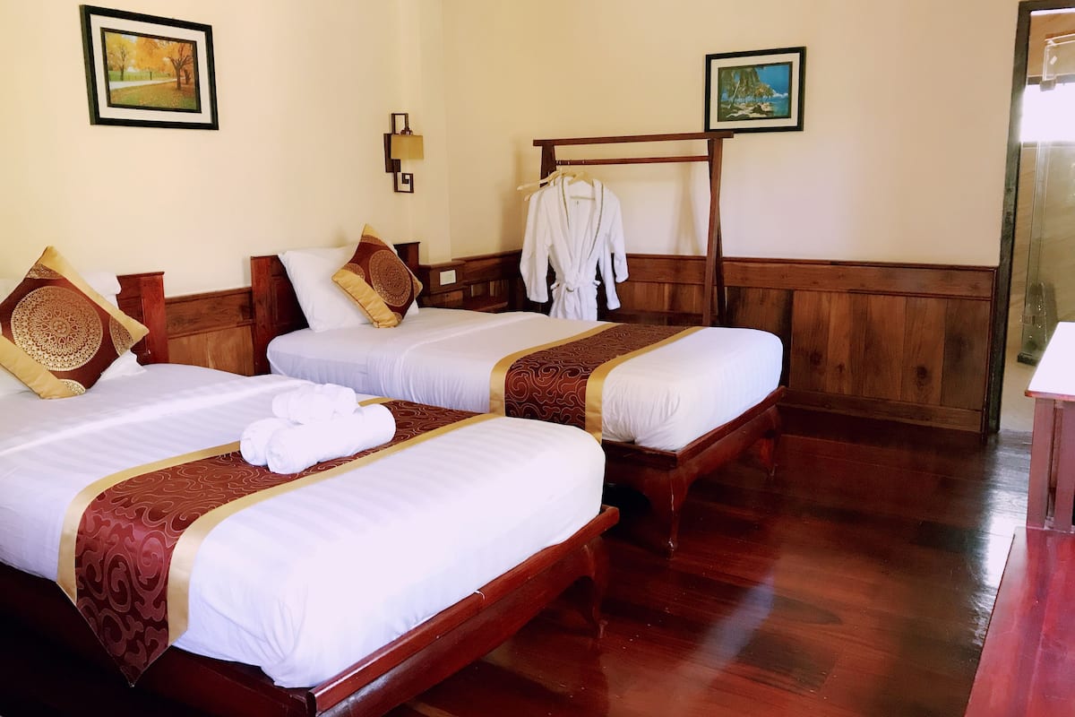 A hotel room with two beds, each with a wooden headboard, white linens, and decorative pillows. There is a wooden bench and a robe rack with a white robe hanging on it.