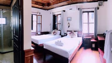 Ein Hotelzimmer mit zwei Einzelbetten, Holzboden, Holzbalkendecke, einem Fenster mit Vorhängen, einer Wand-Klimaanlage und einem kleinen Tisch mit Stuhl.