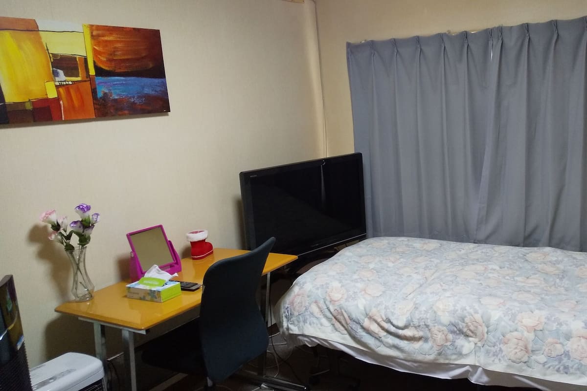 Quarto de hotel com uma cama, uma escrivaninha com computador e uma cadeira, televisão e cortina.