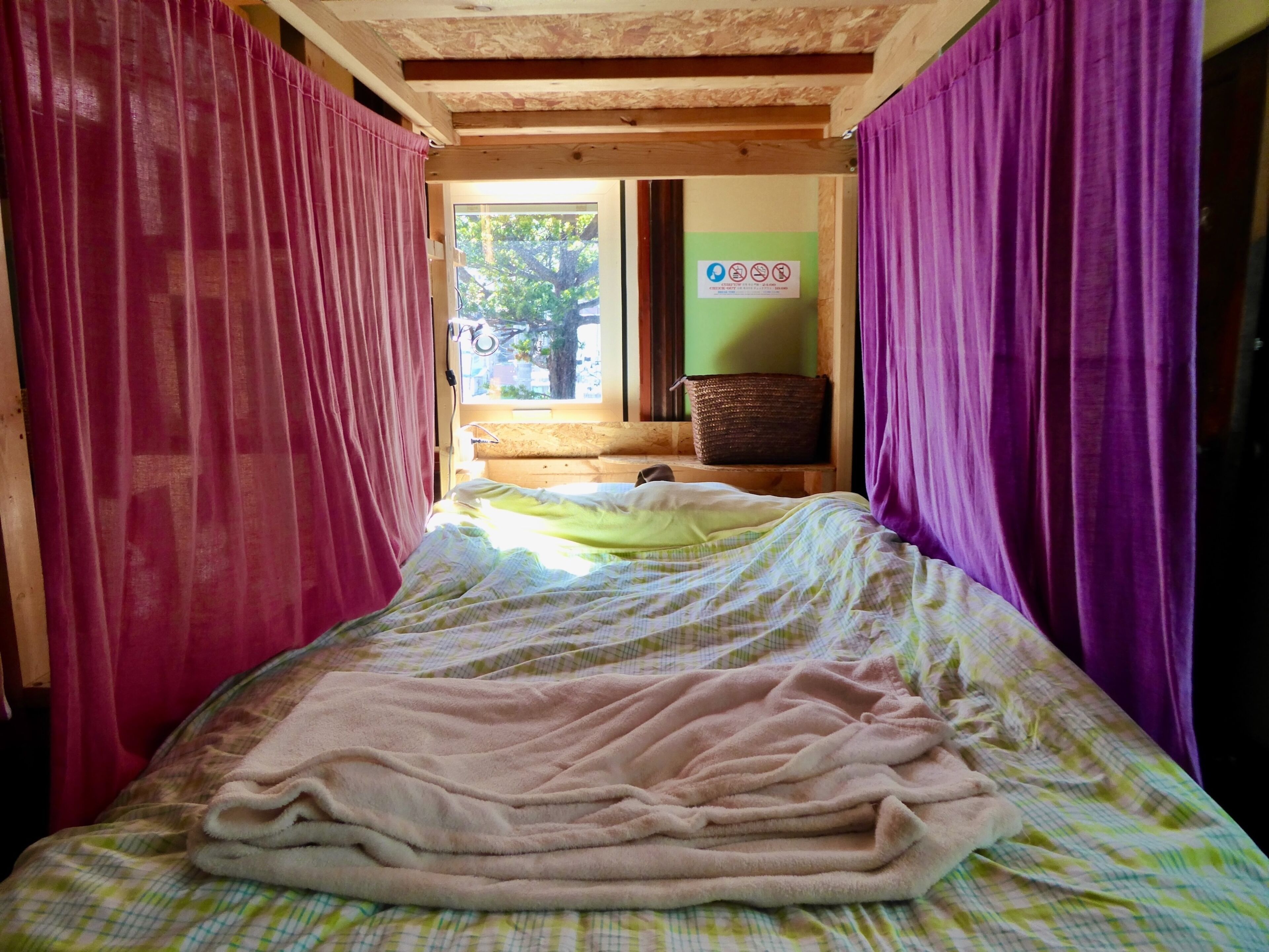 A narrow room with a bed, purple curtains, a window, and a green wall with a sign.
