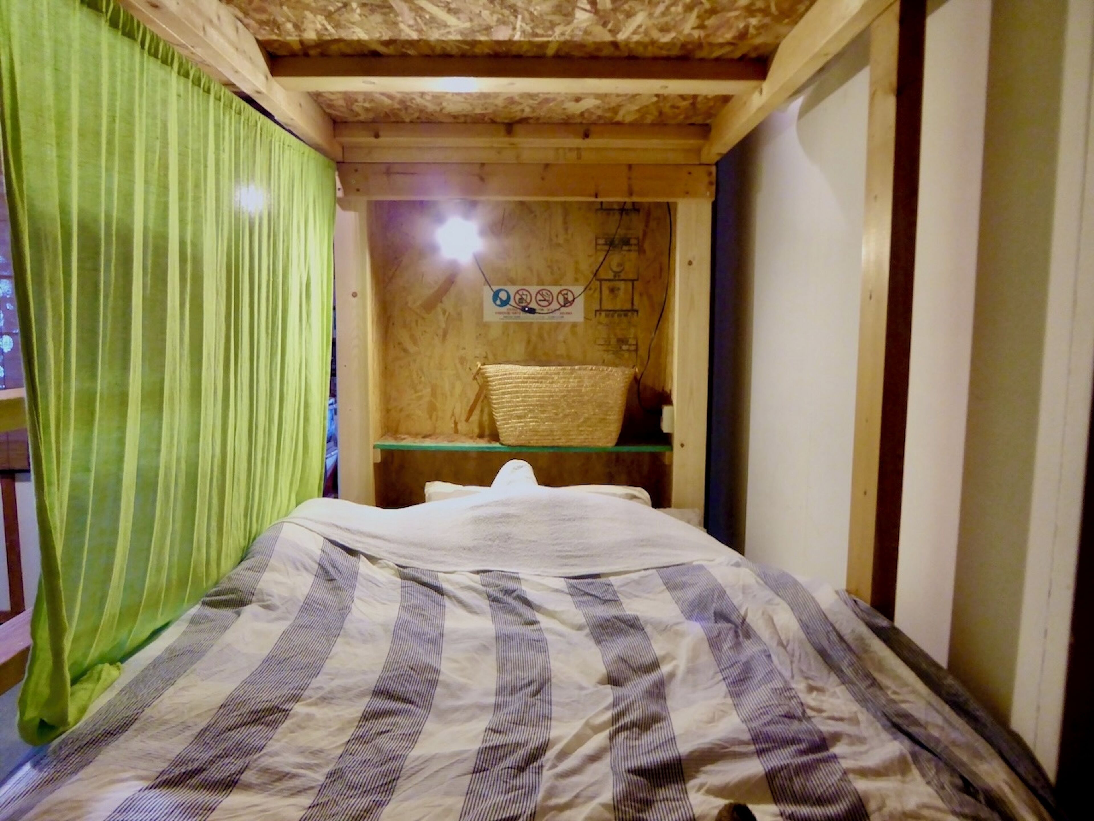 A small, cozy room with a bed, a green curtain, a wooden shelf with a basket, and a sign on the wall.