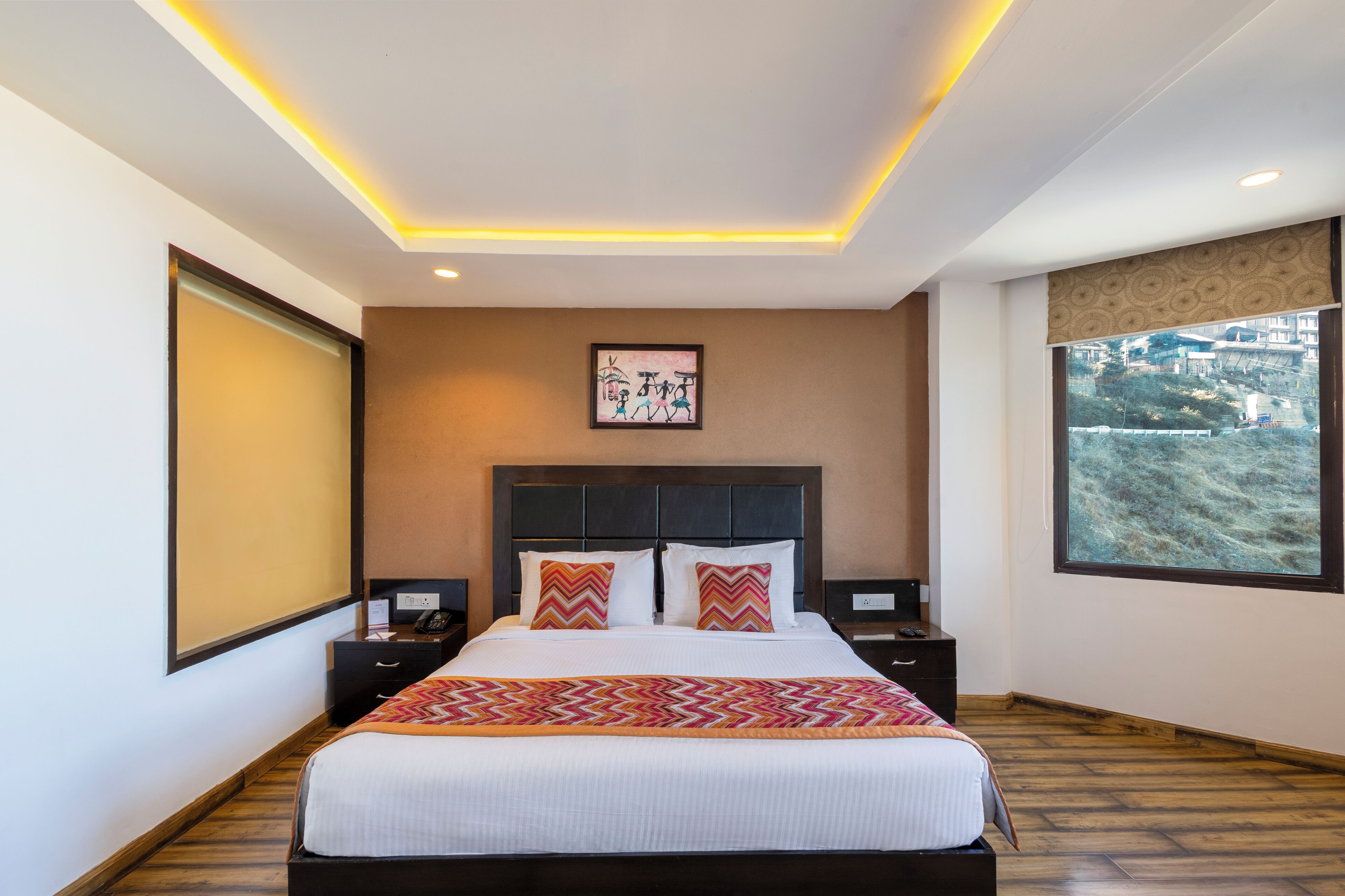 A modern hotel room with a large bed, a headboard with a built-in TV, a bedside table, a nightstand, and a large window with a view.