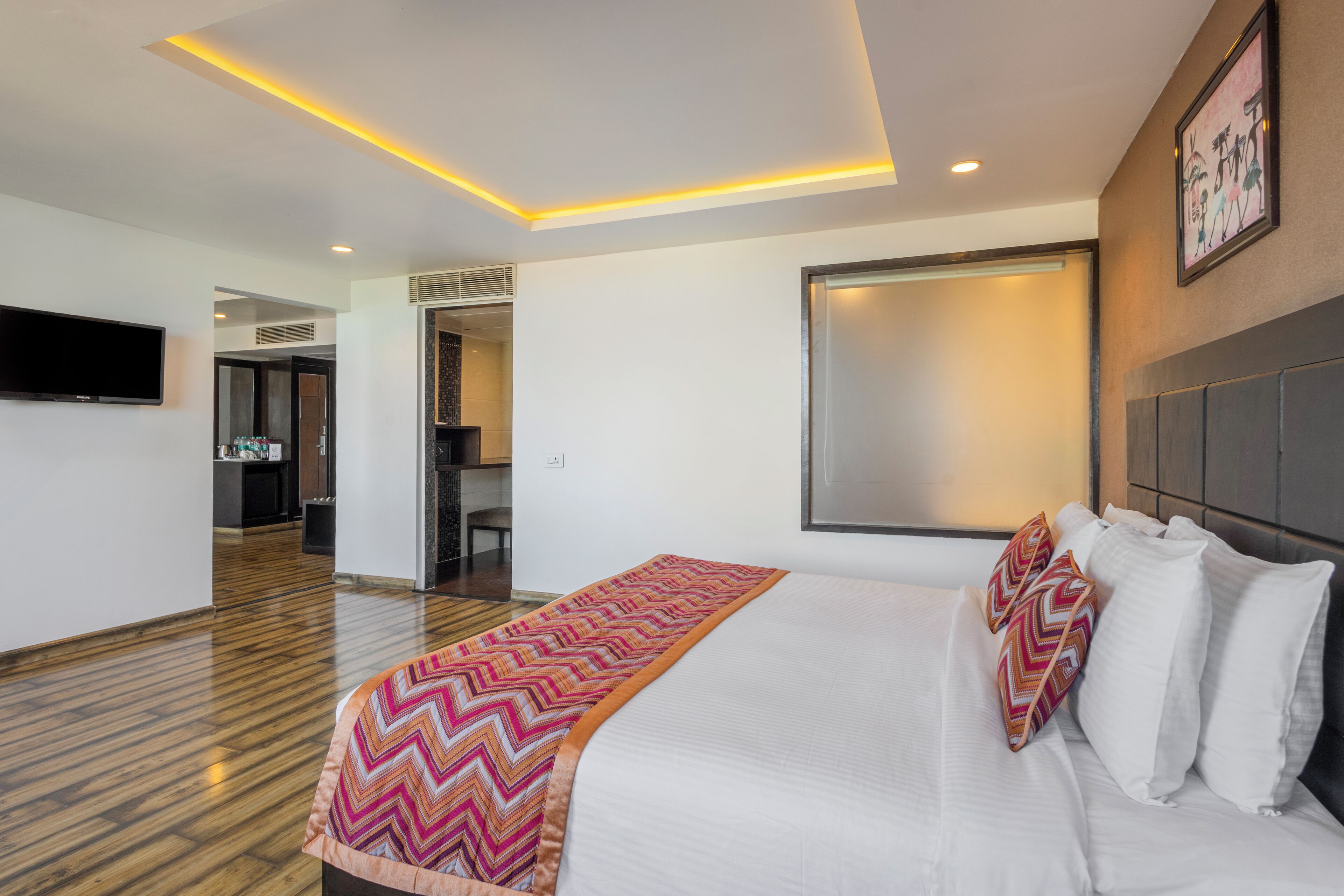 A modern hotel room with a large bed, a flat-screen TV mounted on the wall, and a wooden floor.