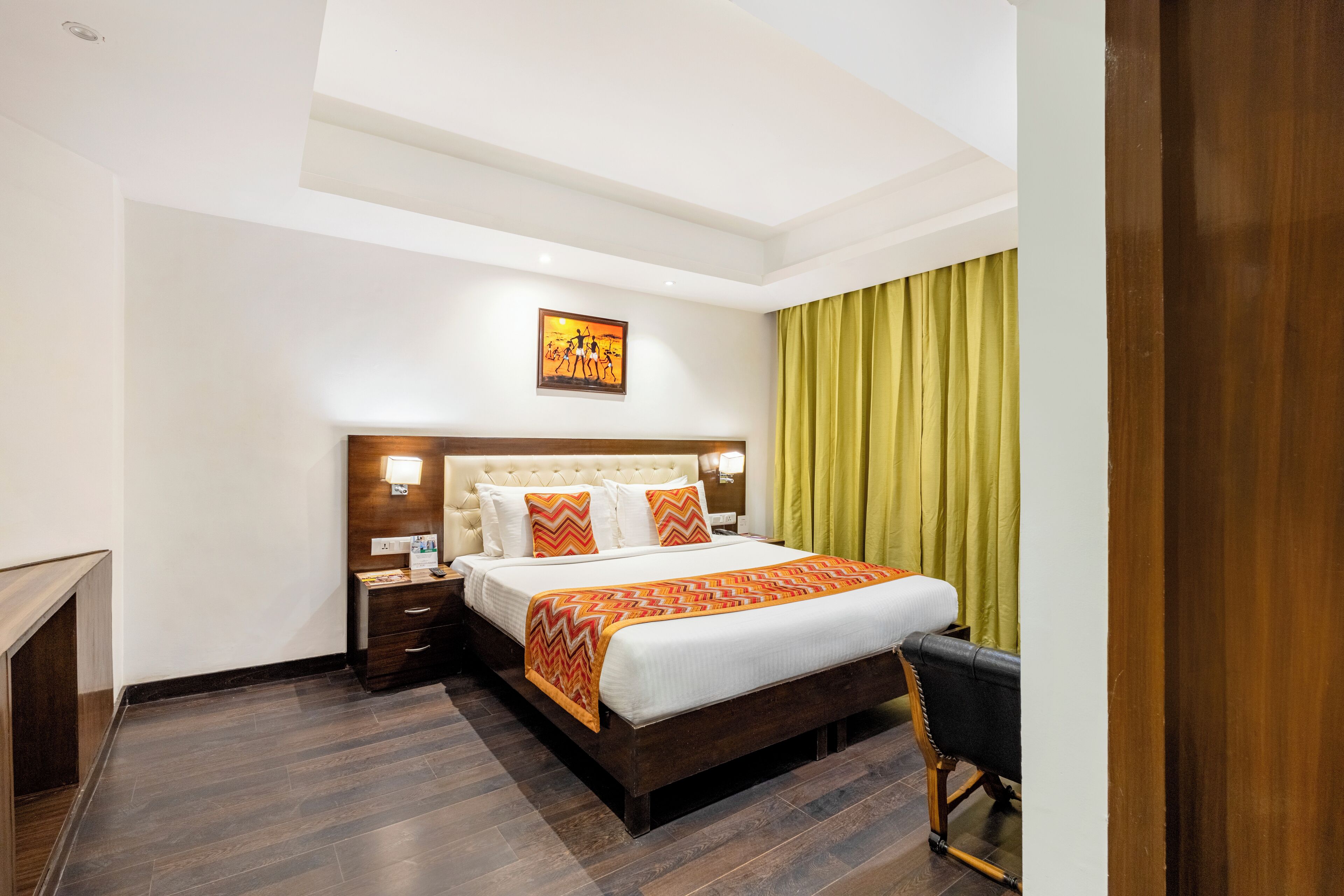 A hotel room with a large bed, a wooden nightstand, a bedside table, a wall-mounted lamp, and a painting above the bed.