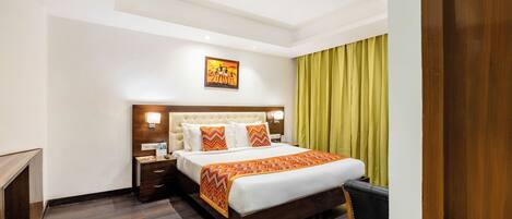 A hotel room with a large bed, a wooden nightstand, a bedside table, a wall-mounted lamp, and a painting above the bed.