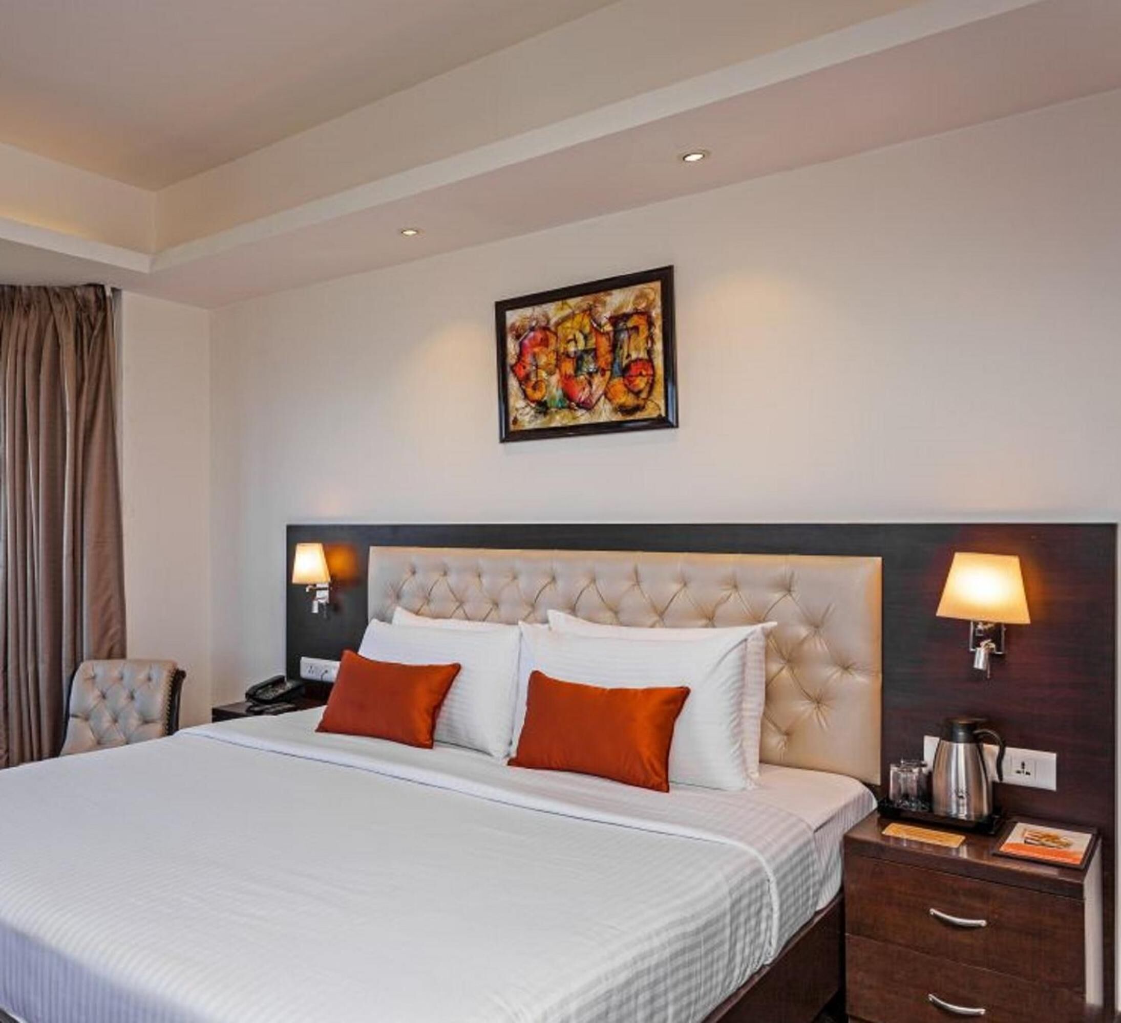 Una habitación de hotel con una cama grande, dos almohadas, cabecera, mesita de noche, lámpara, un cuadro y una silla.