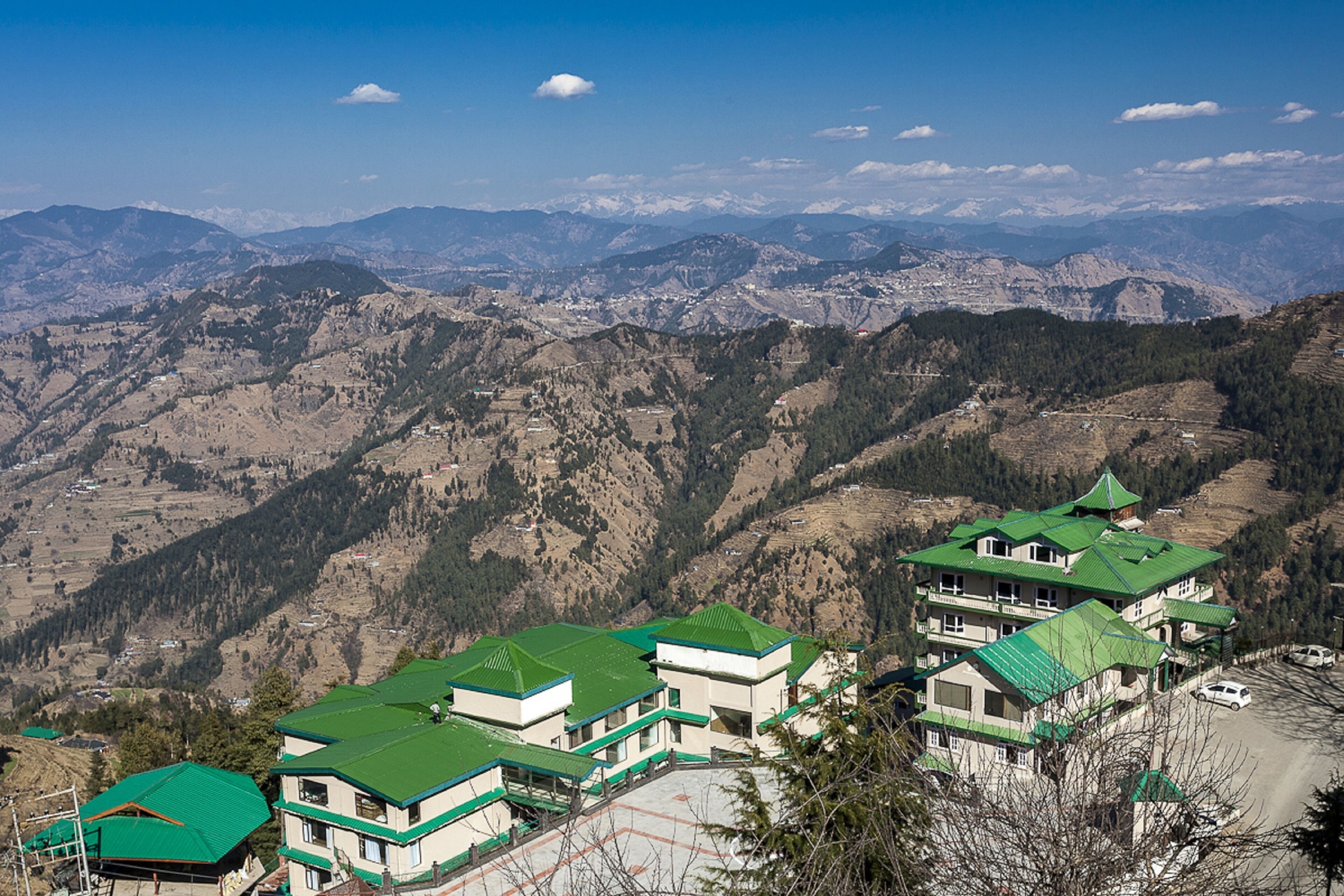 A mountainous landscape with a cluster of buildings featuring green roofs and multiple stories, surrounded by rugged terrain and distant snow-capped peaks.