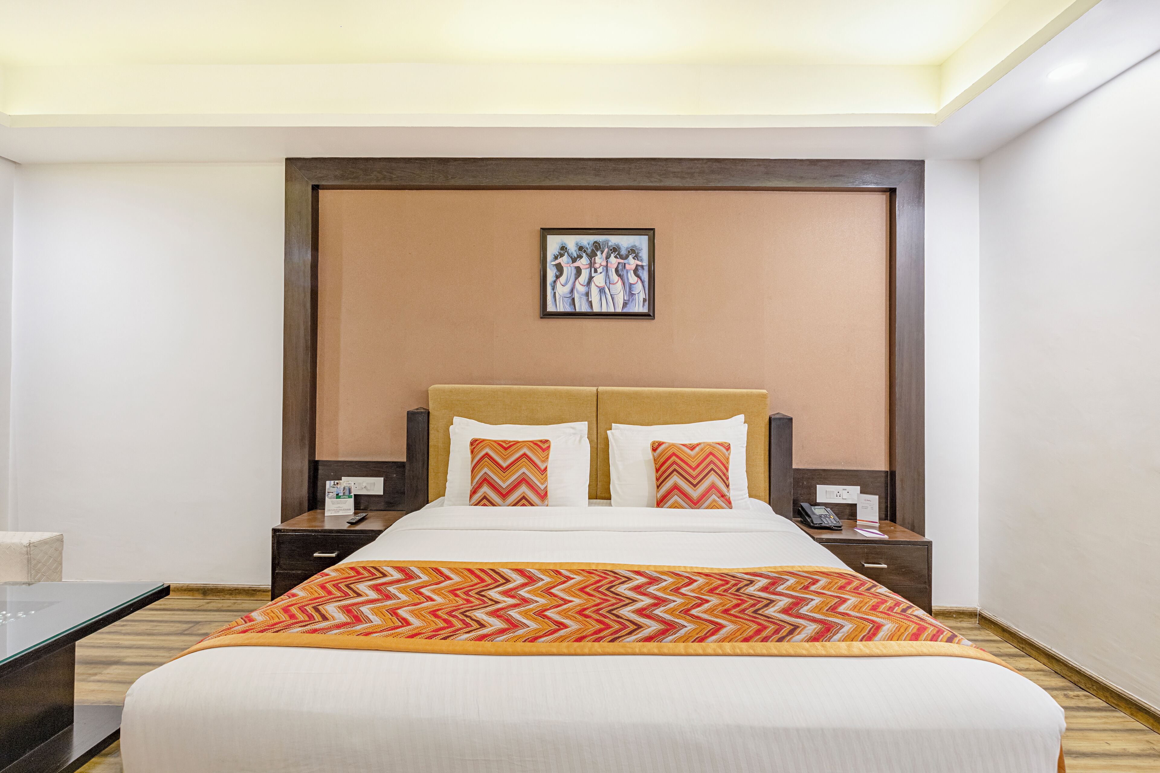 A hotel room with a large bed, two nightstands, a wooden headboard, a framed artwork above the bed, and a patterned throw blanket.