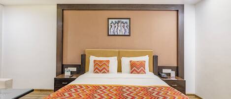 A hotel room with a large bed, two nightstands, a wooden headboard, a framed artwork above the bed, and a patterned throw blanket.