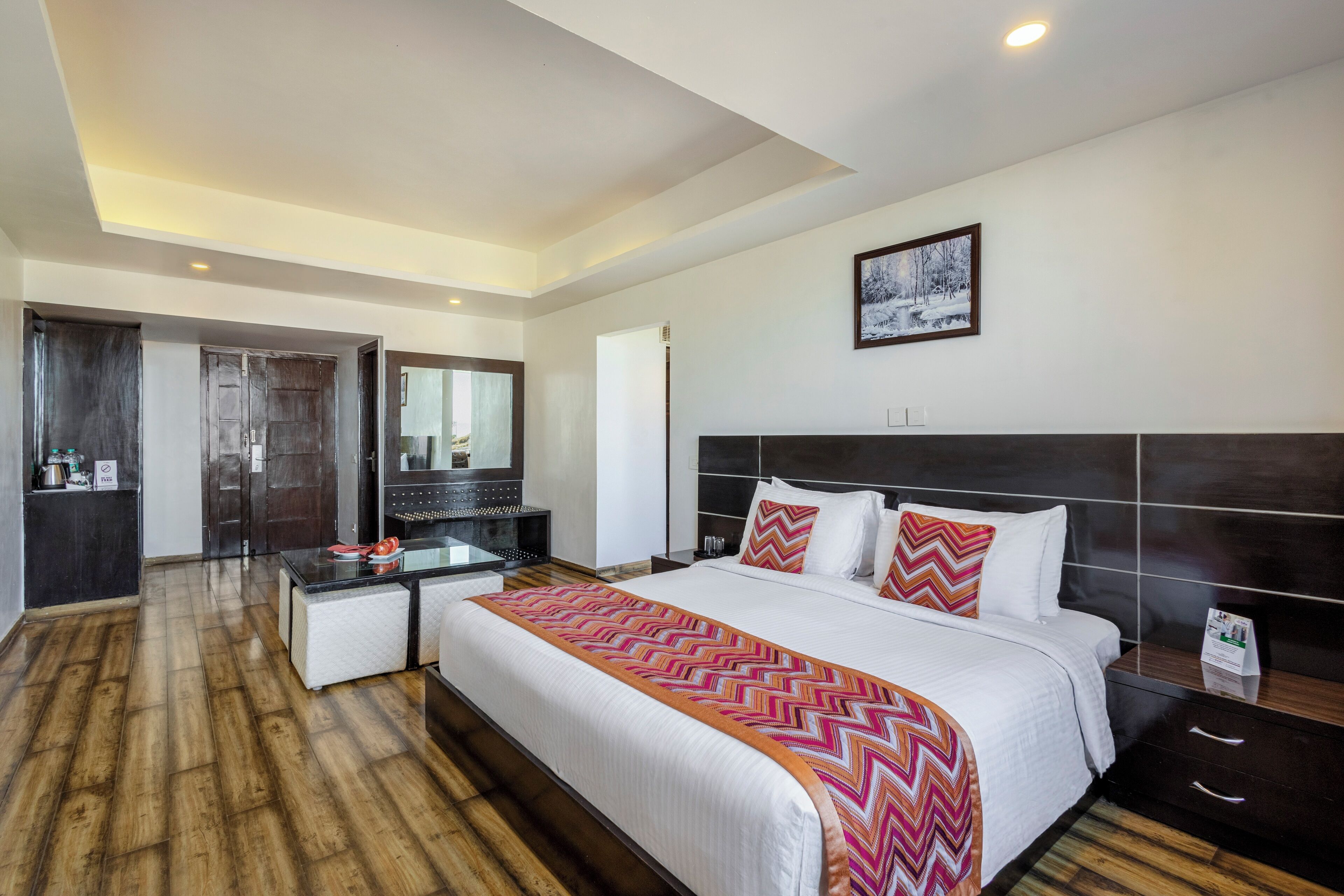 A hotel room with a large bed, a bedside table, a mirror, and a wooden floor.