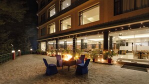 An outdoor seating area with a fire pit and chairs, a building with large windows, and potted plants. - Sterling Kufri (Theog)