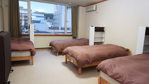 A hotel room with three wooden beds, a desk, a chair, and a window with a view of buildings and mountains. - Otaru Ekimae Guest House Ito - Hostel (Otaru)