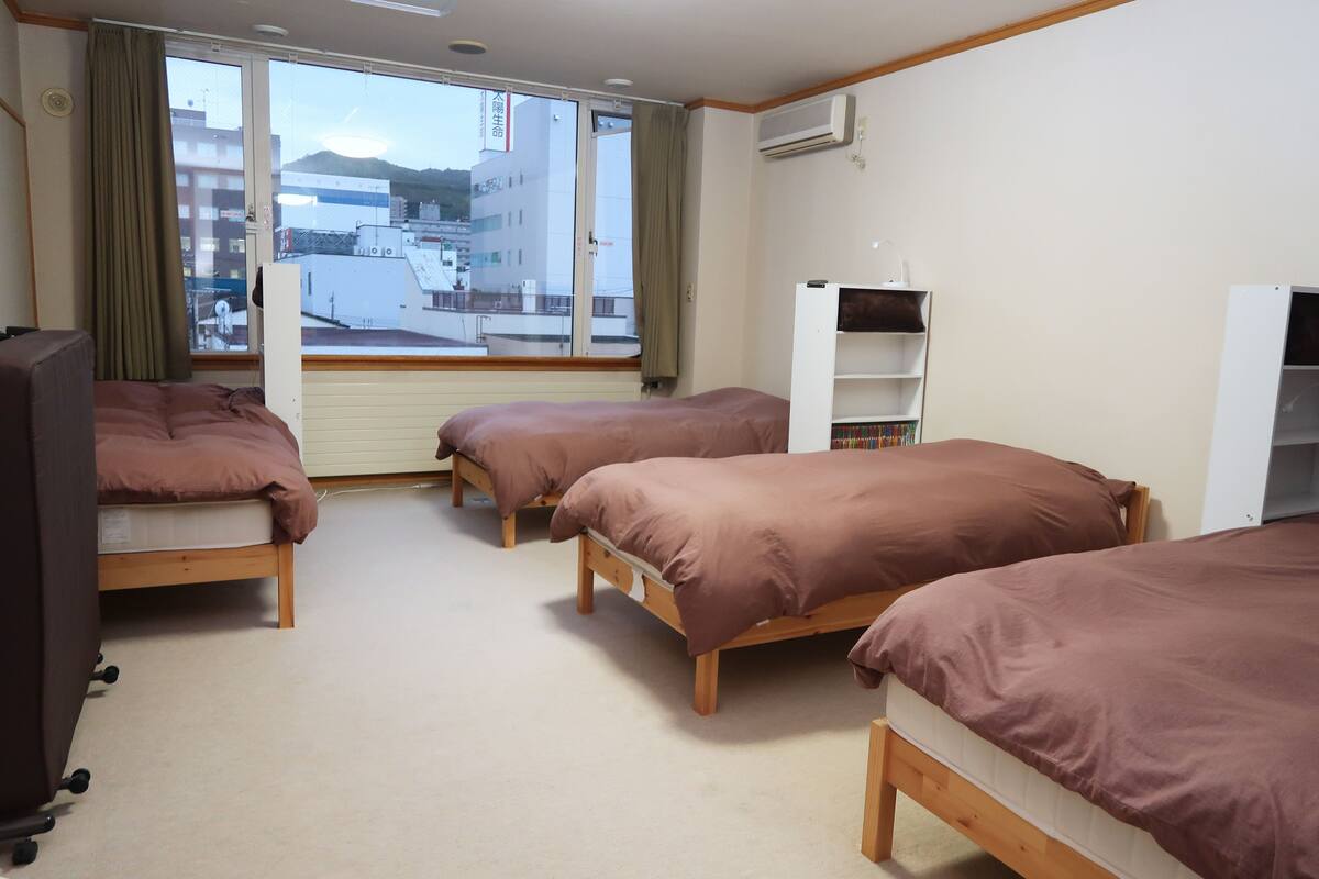 A hotel room with three wooden beds, a desk, a chair, and a window with a view of buildings and mountains.