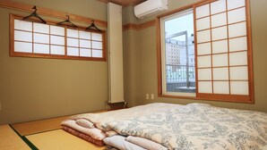 A traditional Japanese-style room with tatami flooring, a futon bed, sliding shoji doors, and a view of buildings outside. - Otaru Ekimae Guest House Ito - Hostel (Otaru)