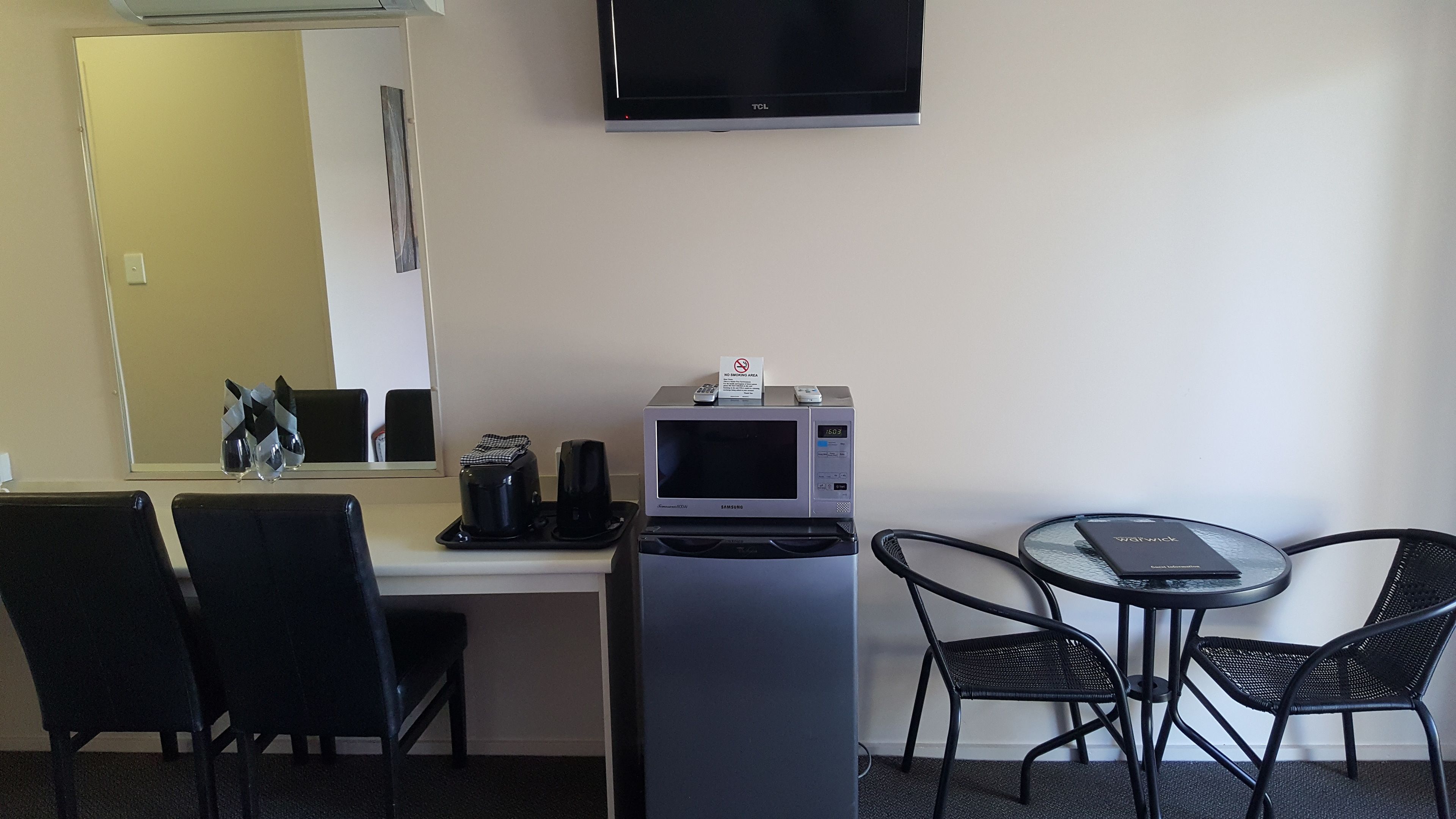 Habitación de hotel con escritorio, dos sillas, microondas, refrigerador pequeño, televisor montado en la pared y una mesa redonda con una computadora portátil.