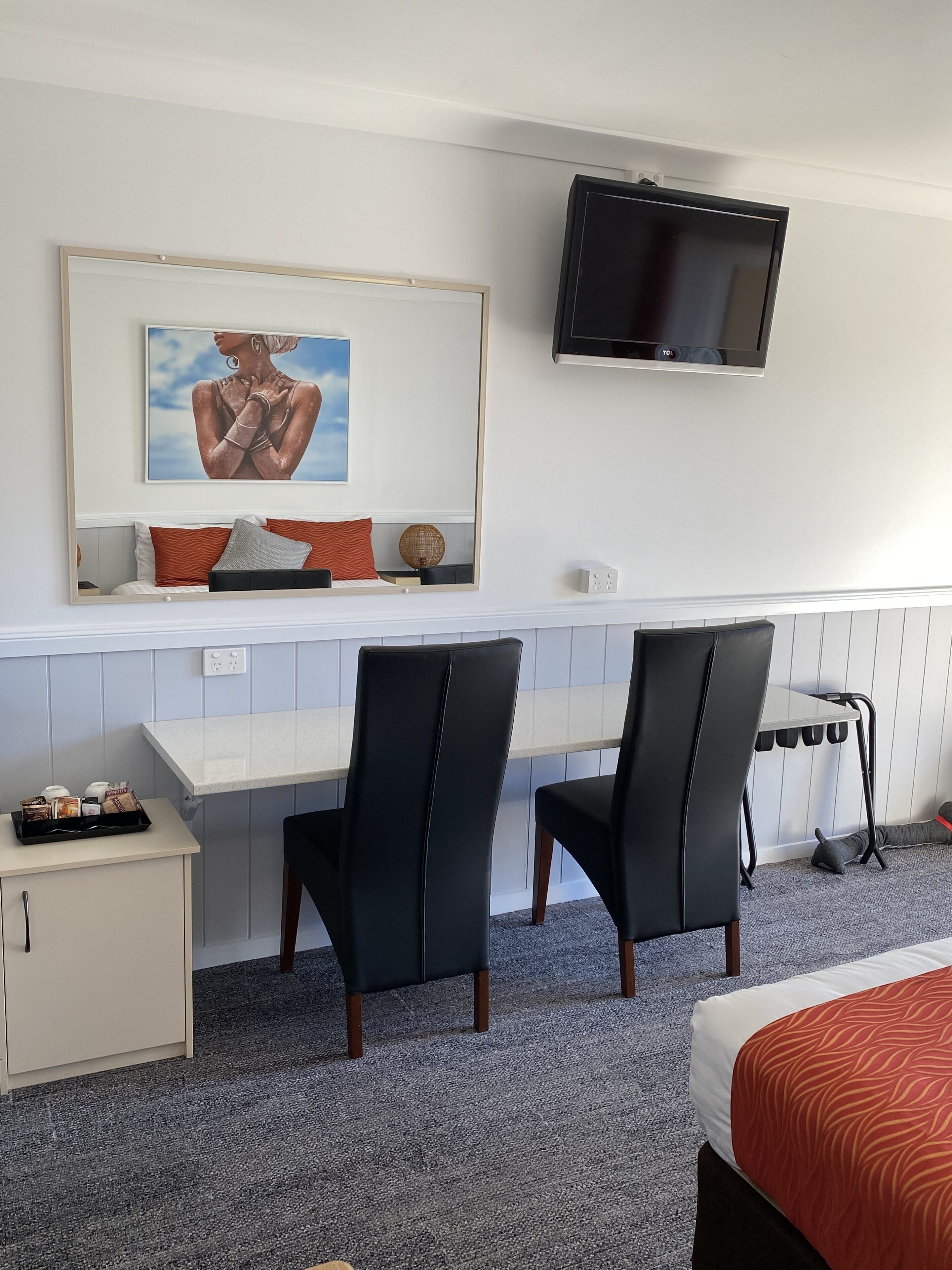 A hotel room with a bed, a desk, two chairs, a TV, a painting, and a small cabinet.