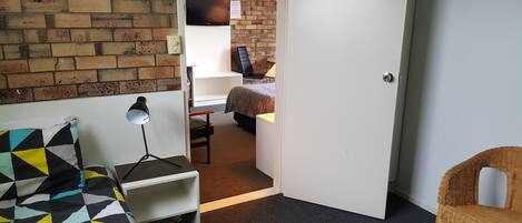A room with a brick wall, a bed, a desk with a lamp, and a wicker chair.