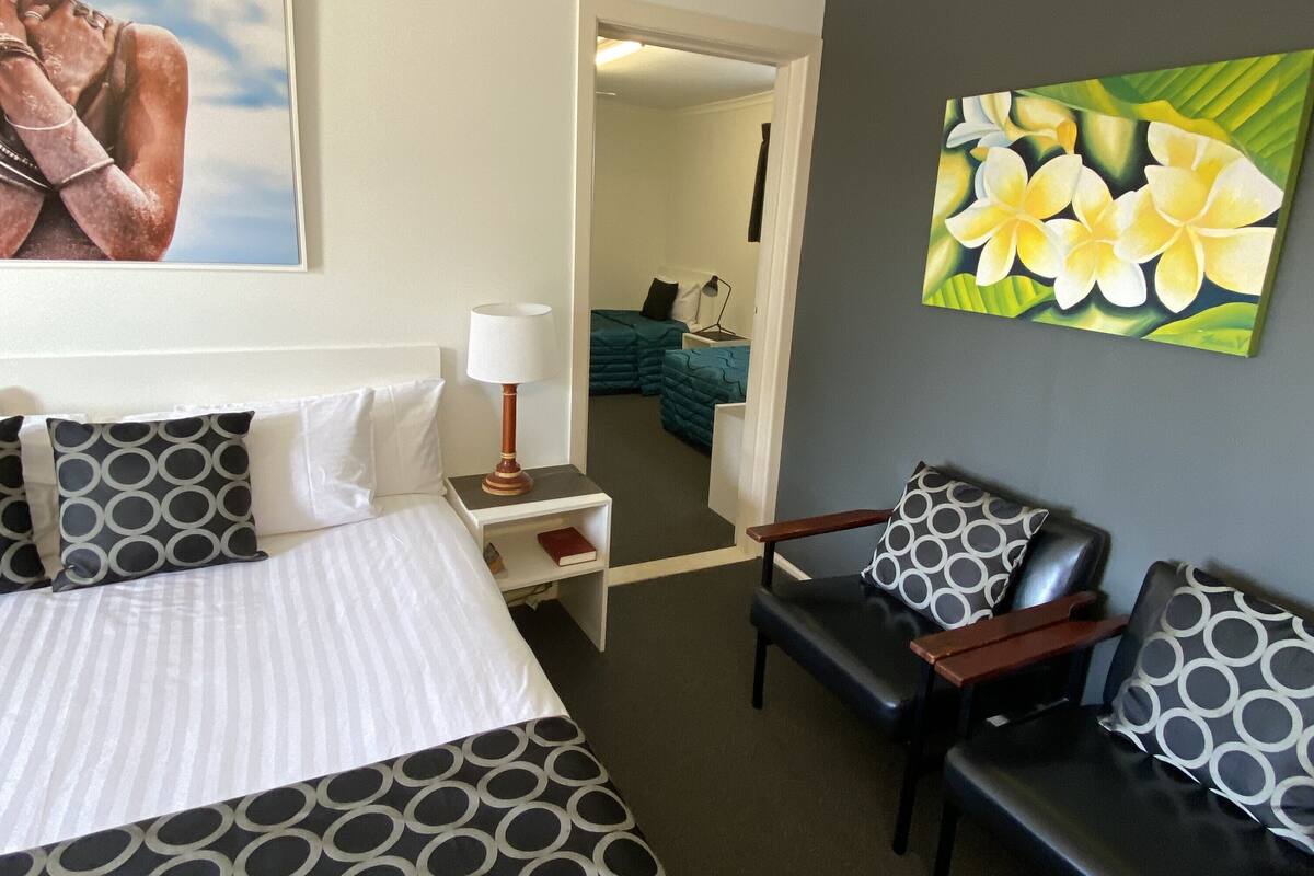 A hotel room with a bed, two chairs, a nightstand, a lamp, a painting of flowers, and a view into the next room.