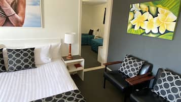 A hotel room with a bed, two chairs, a nightstand, a lamp, a painting of flowers, and a view into the next room.