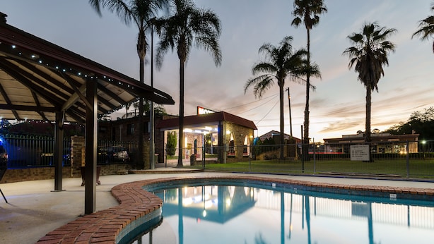 Piscina con borde de ladrillo, rodeada de palmeras y una zona cubierta con luces.
