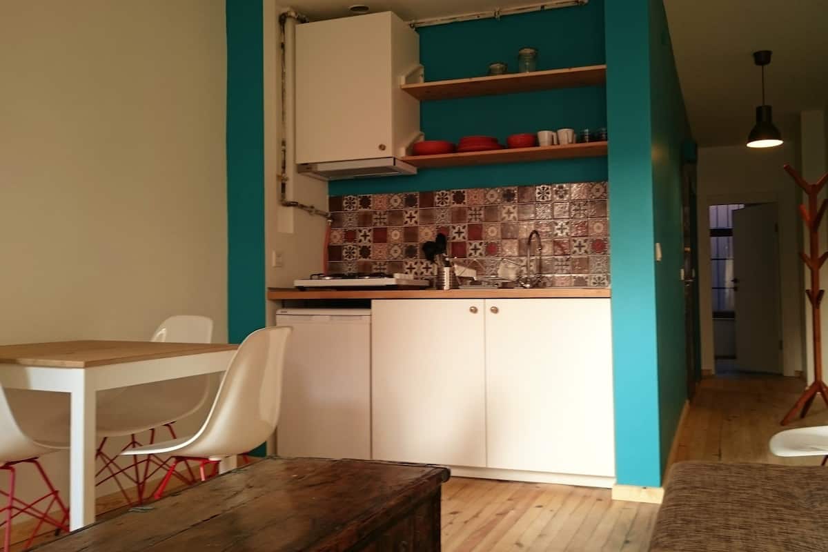 A compact kitchen with white cabinets, a tiled backsplash, and a wooden countertop. There is a dining table with two chairs and a bench.