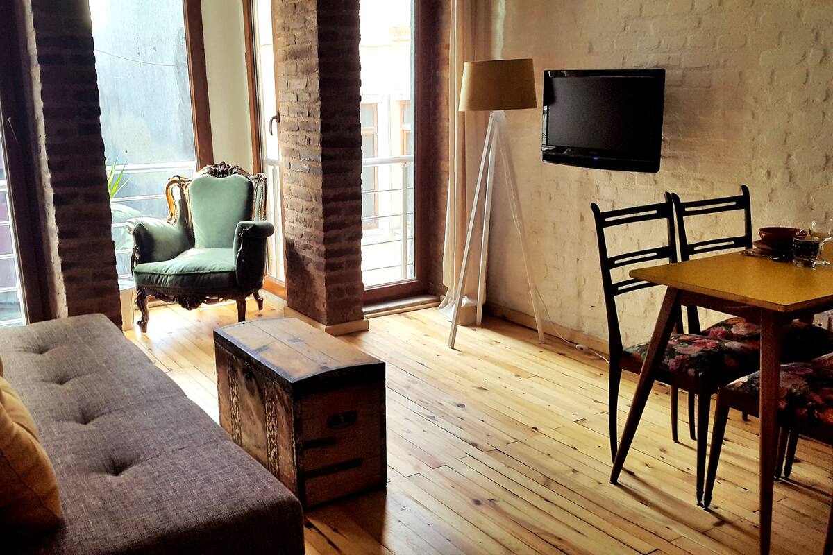 A cozy room with a wooden floor, a green armchair, a sofa, a dining table with chairs, and a TV mounted on the wall.
