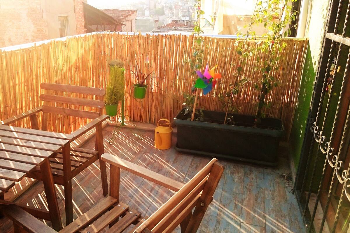 A wooden table and chairs on a balcony with a bamboo fence, potted plants, and a black planter.