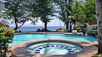 A pool with a hot tub, surrounded by tropical plants and a view of the ocean.