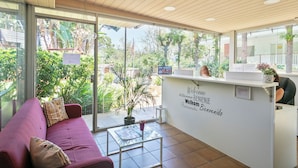 A reception area with a wooden ceiling, a reception desk, a pink sofa, and a glass wall offering a view of greenery outside.