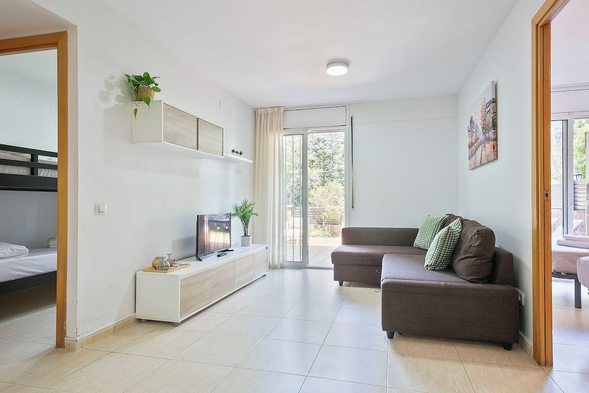 A living room with a sofa, a TV on a stand, a plant, and a sliding door leading to an outdoor area.