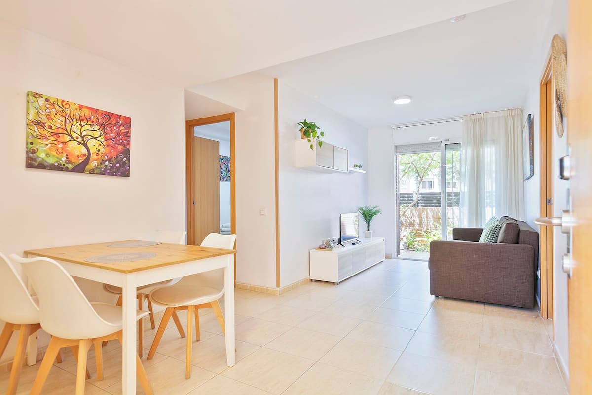 A modern living room with a sofa, a dining table with chairs, a painting on the wall, and a sliding glass door leading to an outdoor area.