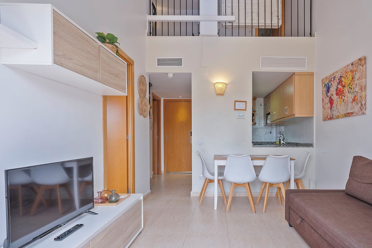 A modern apartment with a TV, dining area, kitchenette, and a balcony.