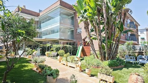 A building with a glass facade, a well-maintained garden with various plants and a bench, and a clear sky.