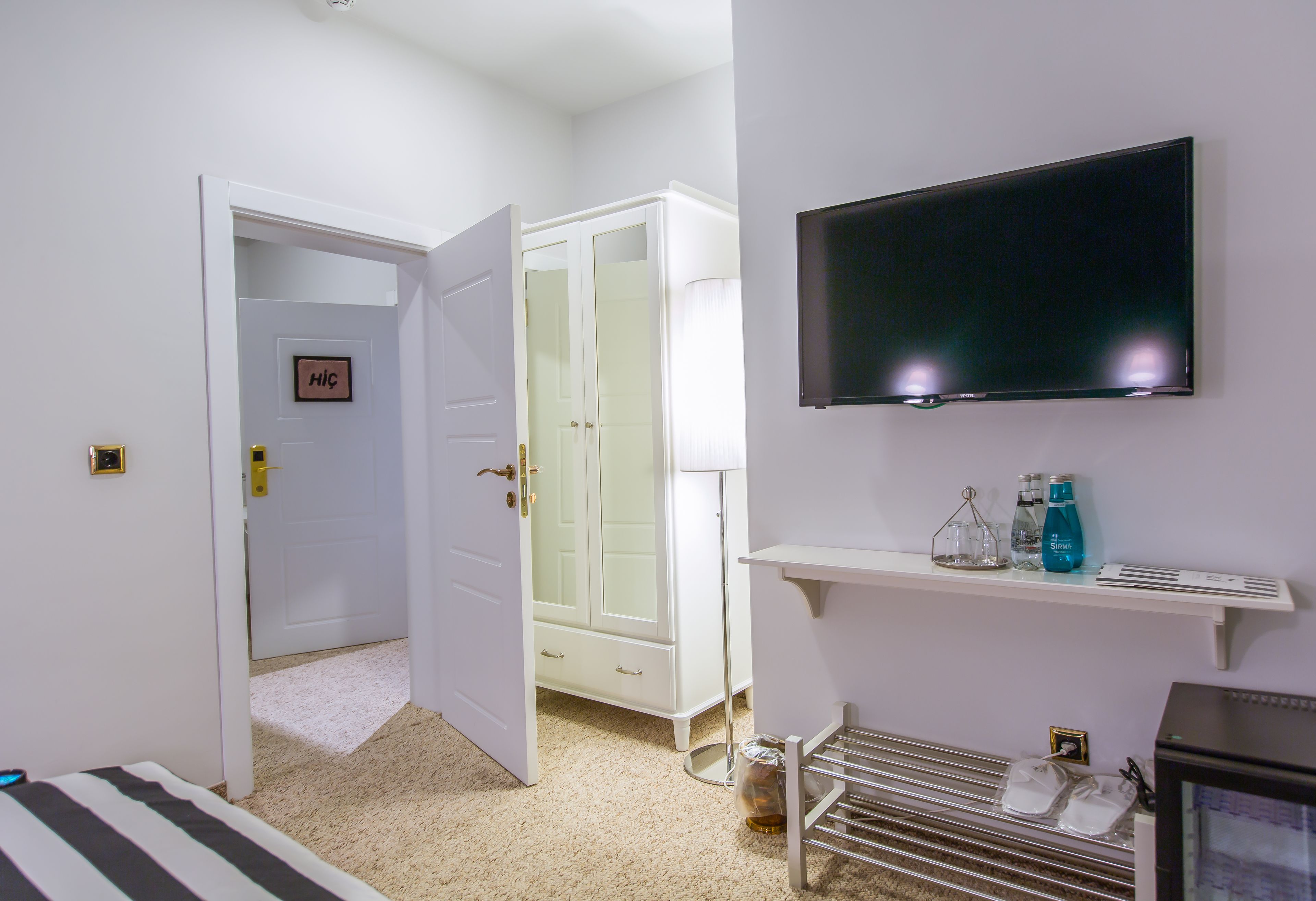 A hotel room with a bed, a television mounted on the wall, a wardrobe, and a small shelf with toiletries.