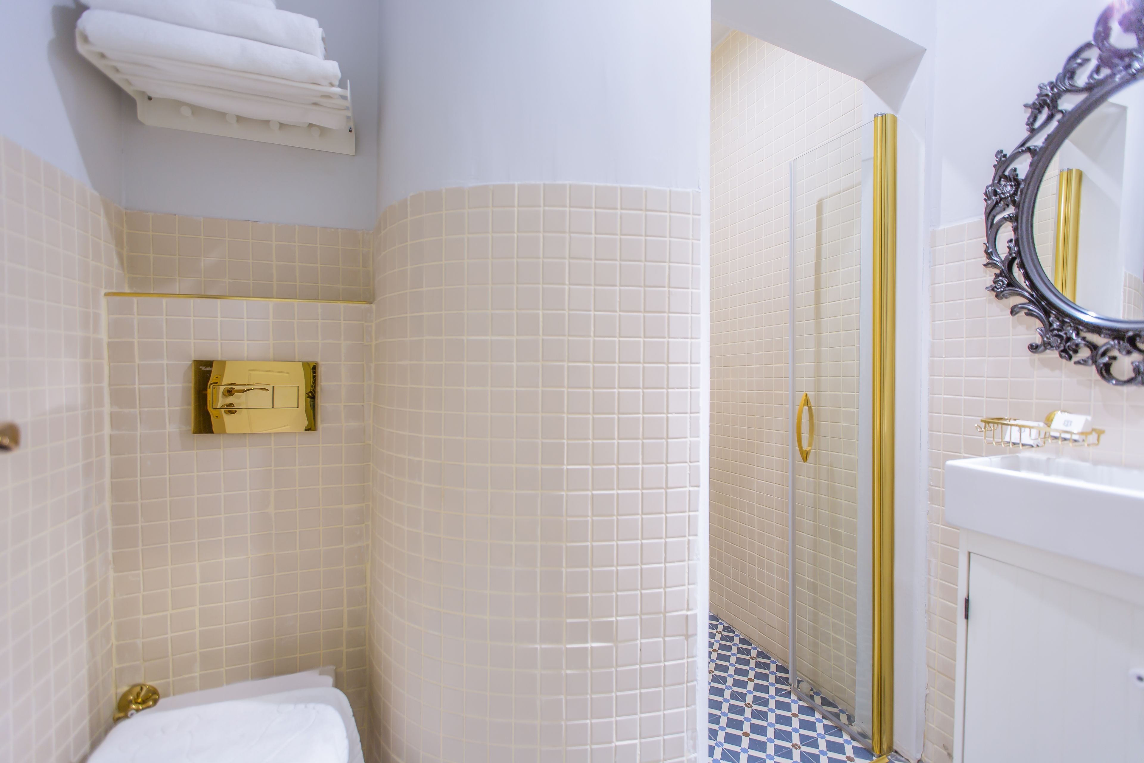 A bathroom with white tiled walls, a gold-tiled shower area, a white sink with gold fixtures, and a mirror with an ornate frame.