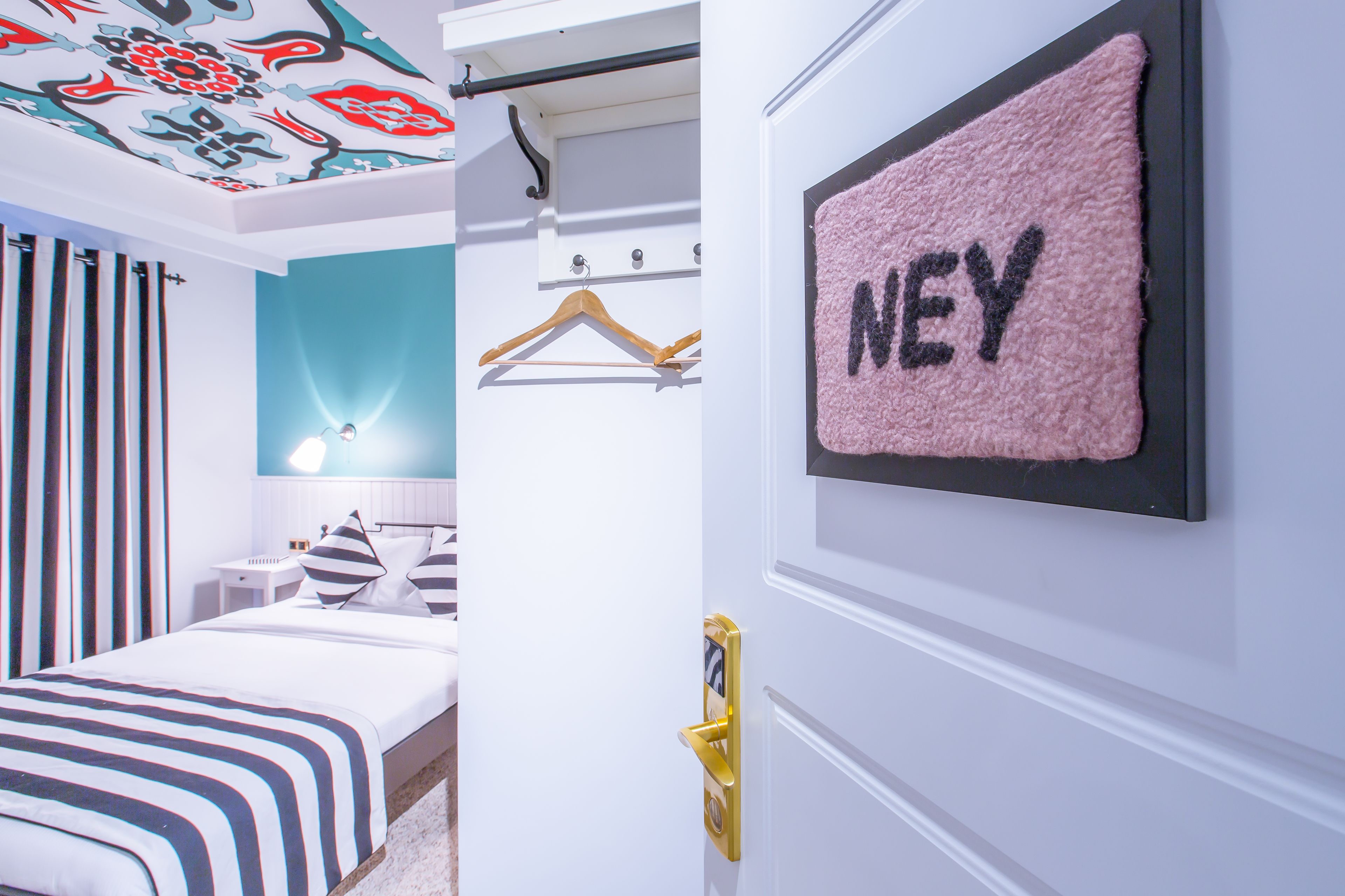 A bedroom with a bed, a bedside table, a hanging light, a wall-mounted towel rack with the name "NEY", and a door with a golden handle.