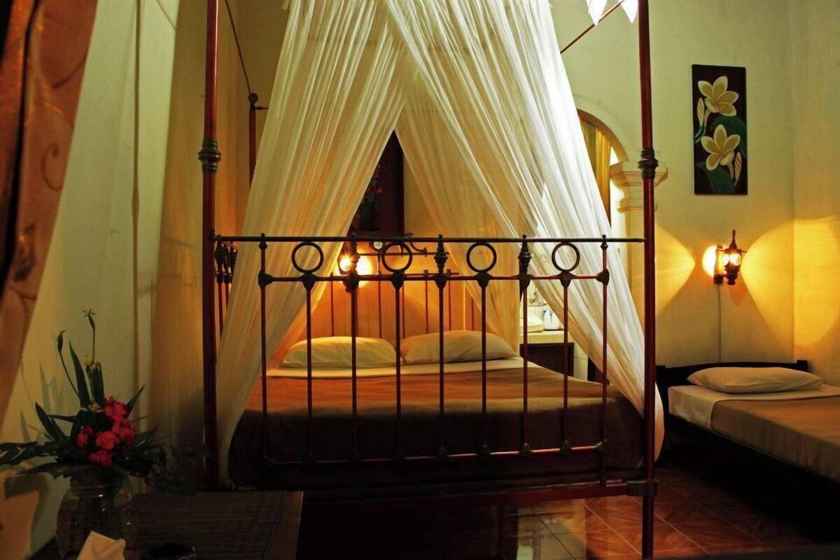 A room with a bed under a canopy, a small table with a vase, and a wall-mounted light fixture.