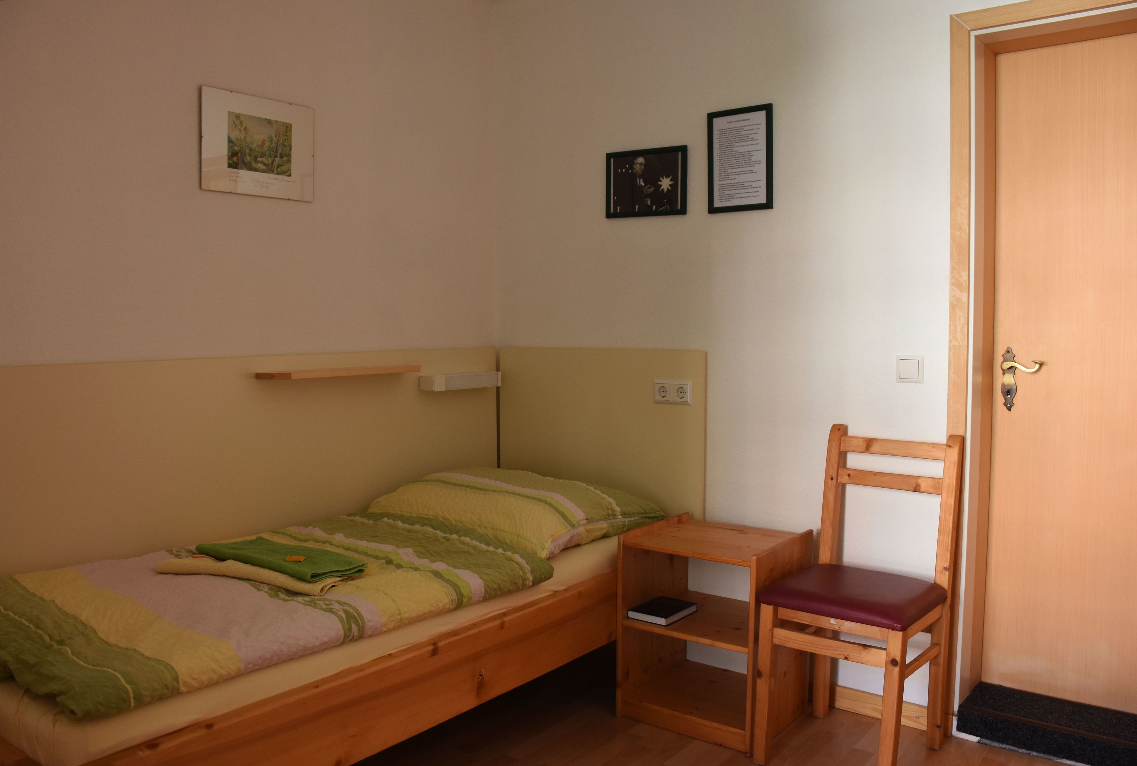 A small, single-bed room with a wooden headboard, a wooden chair, a bedside table, and two framed pictures on the wall.