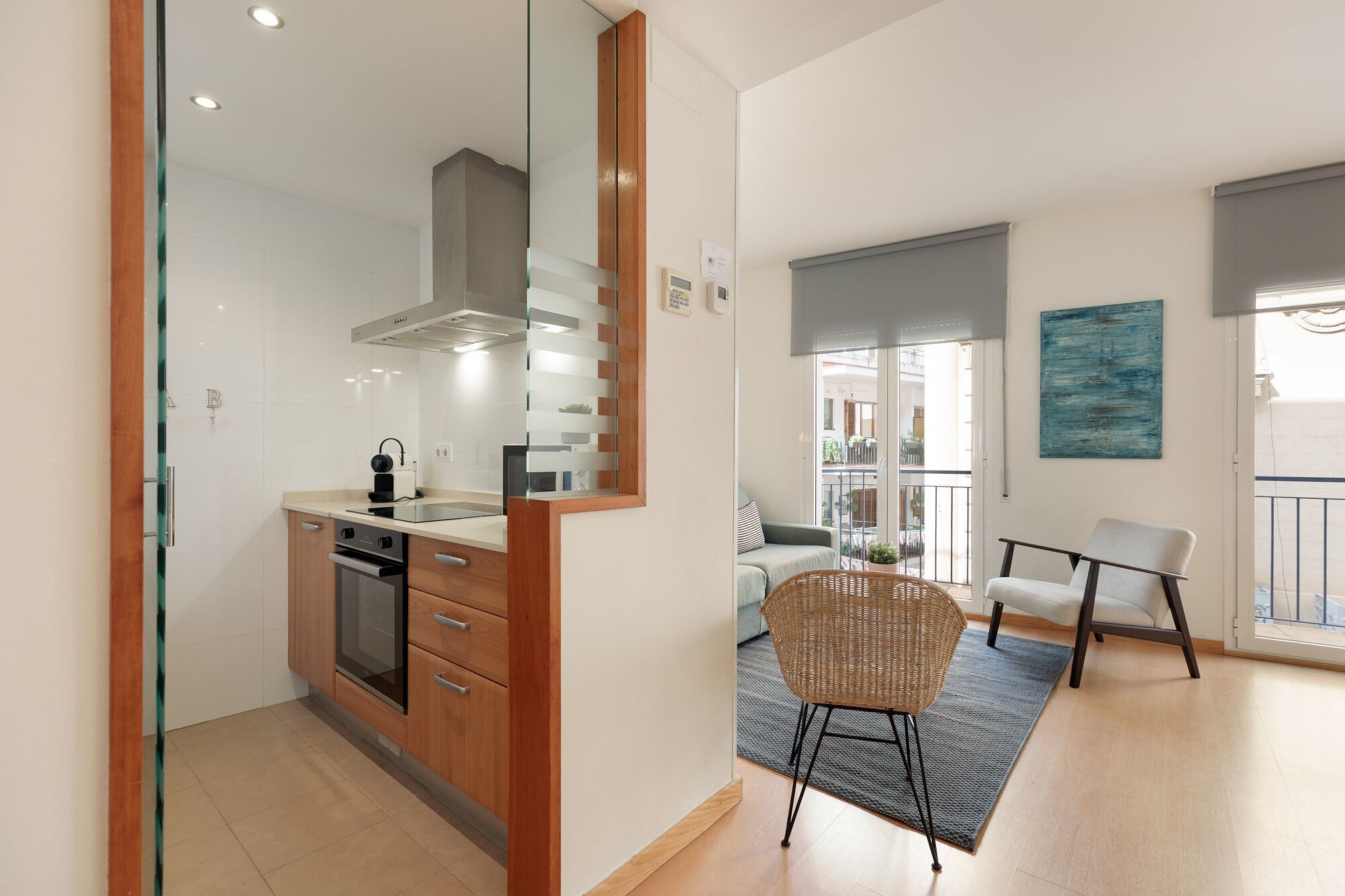 Una cocina moderna con horno empotrado, microondas y cafetera. Hay una zona de comedor con una silla de mimbre y un sofá.