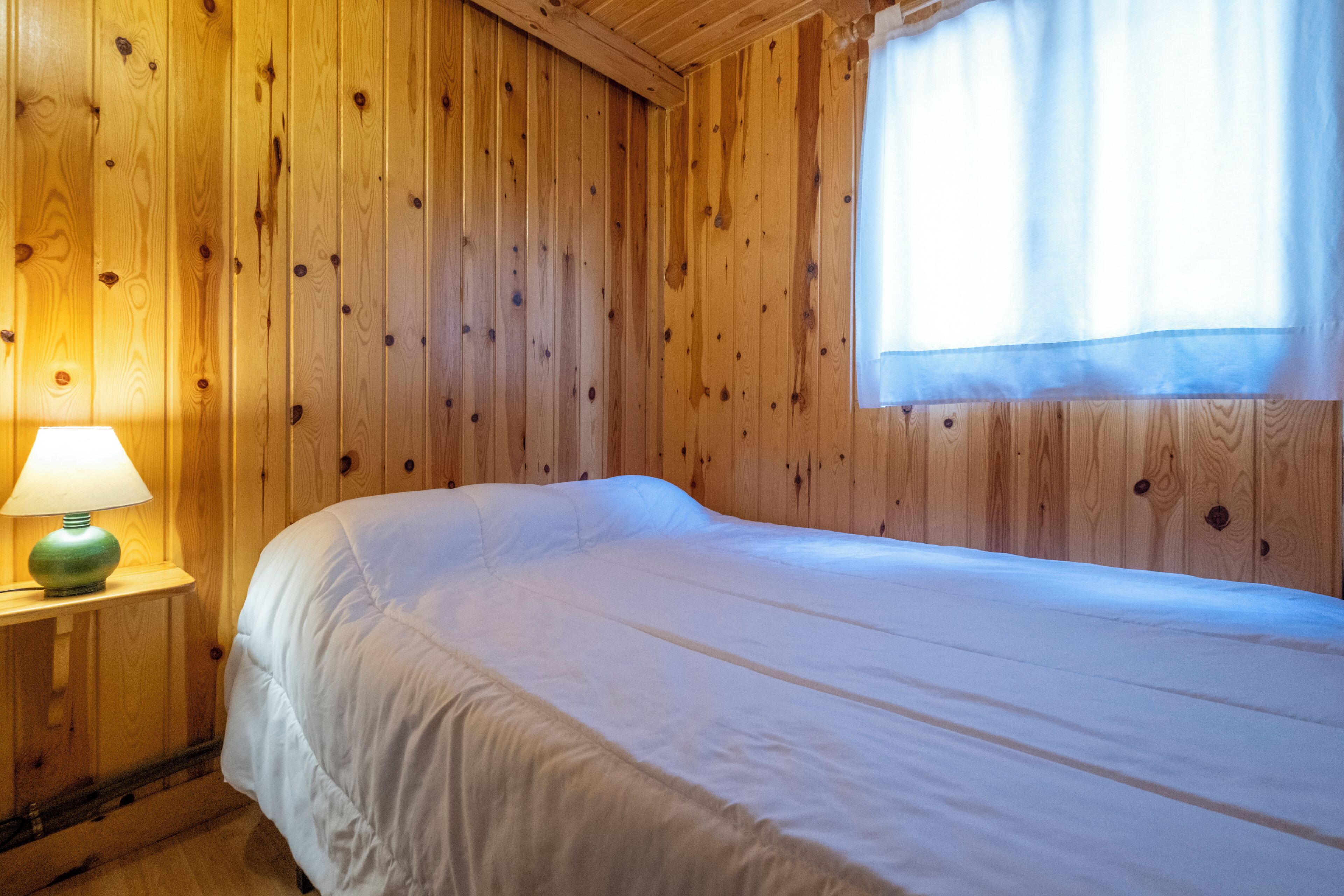 Een kamer met houten lambrisering, een bed, een nachtkastje met lamp en een raam met gordijnen.