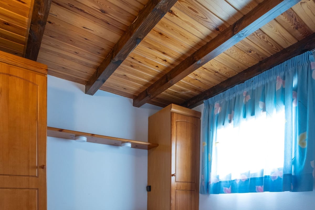 Un dormitorio con techo de madera, cama con ropa de cama blanca, armario de madera y ventana con cortinas azules.
