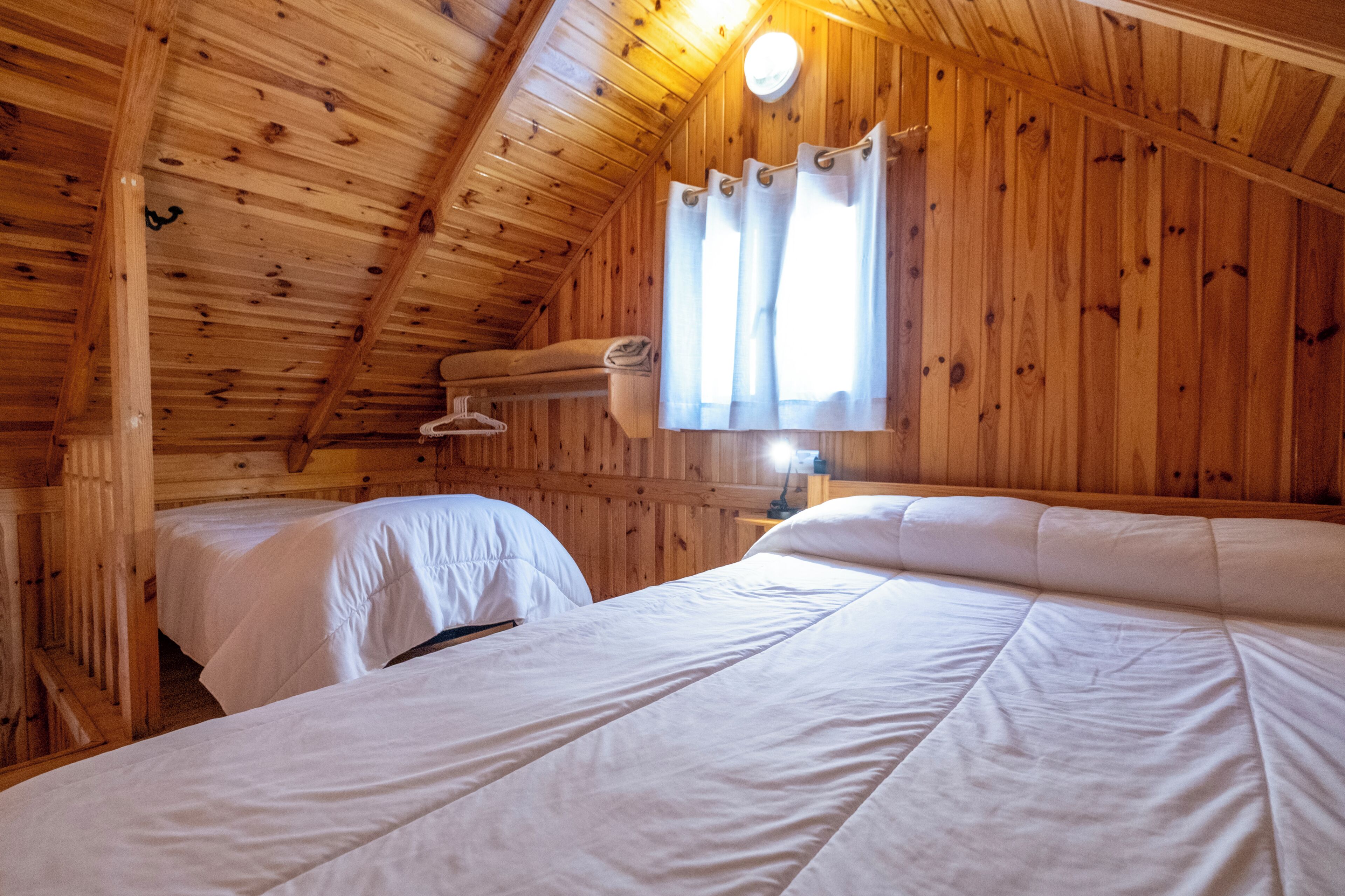 Eine Holzhütte mit geneigter Decke, ausgestattet mit zwei Betten mit weißer Bettwäsche und einem Fenster mit Vorhängen.