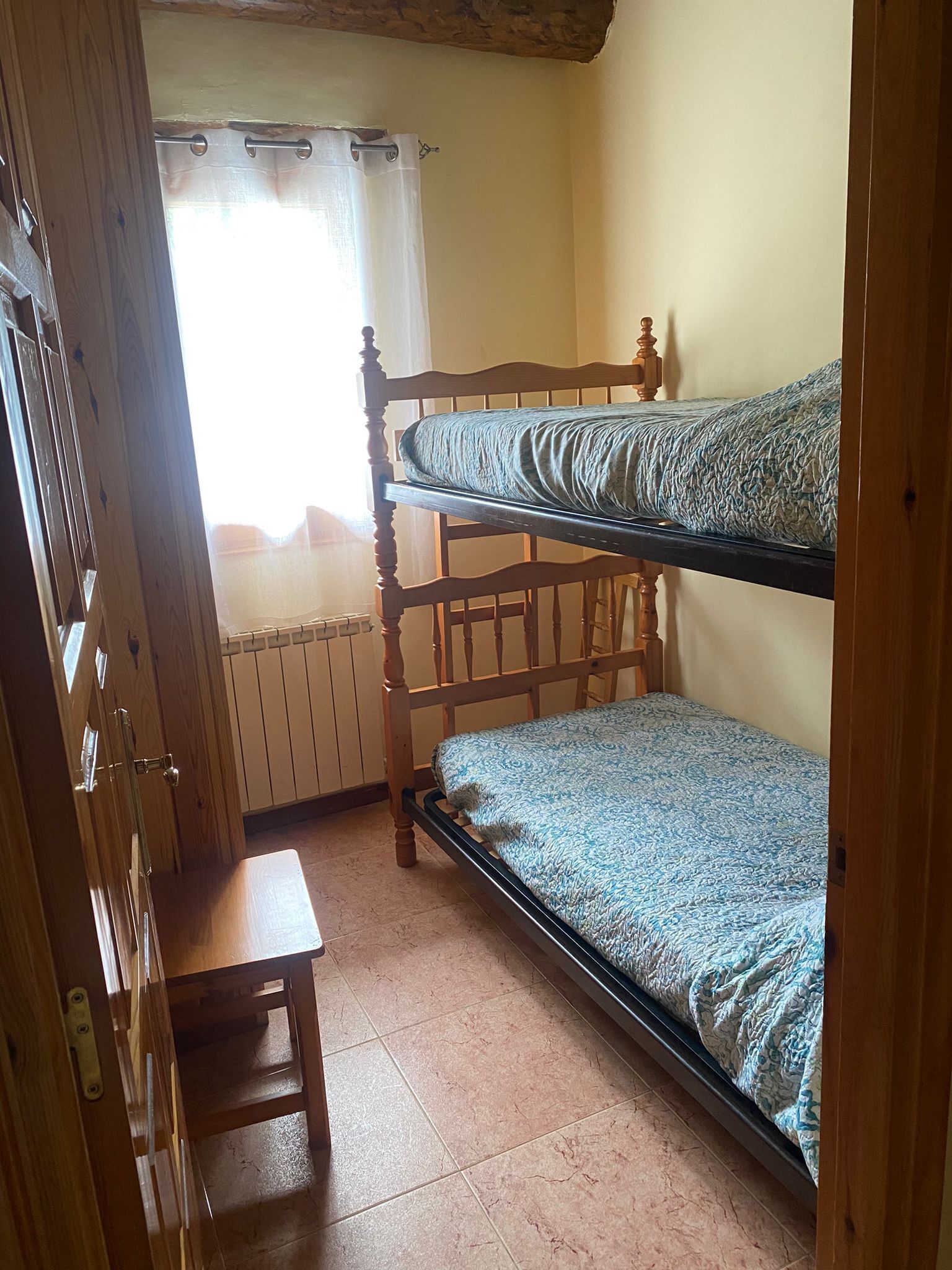 A bunk bed room with a wooden floor, a small wooden table, and a window with curtains.