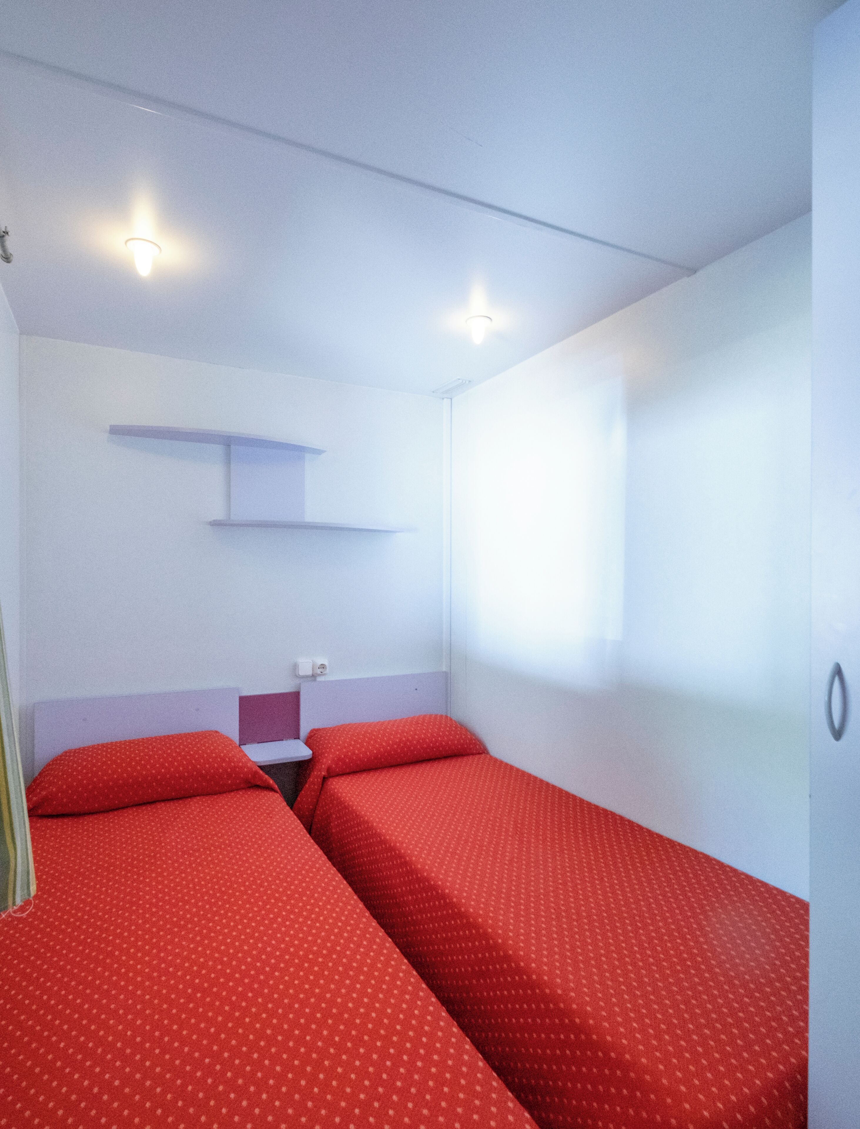 Una habitación pequeña con dos camas, un estante y una ventana.