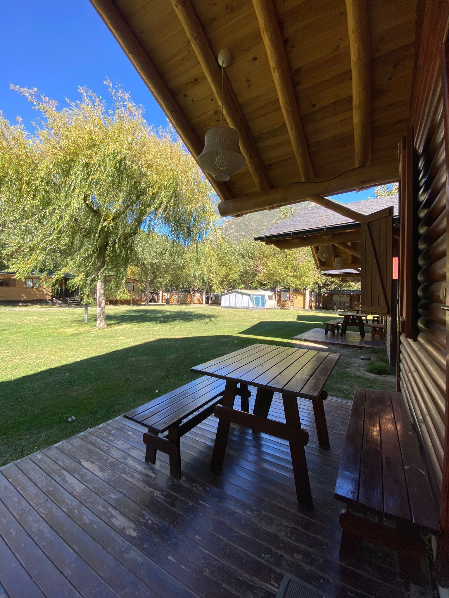 Una terraza de madera con mesas de picnic, un área cubierta y un césped con árboles.