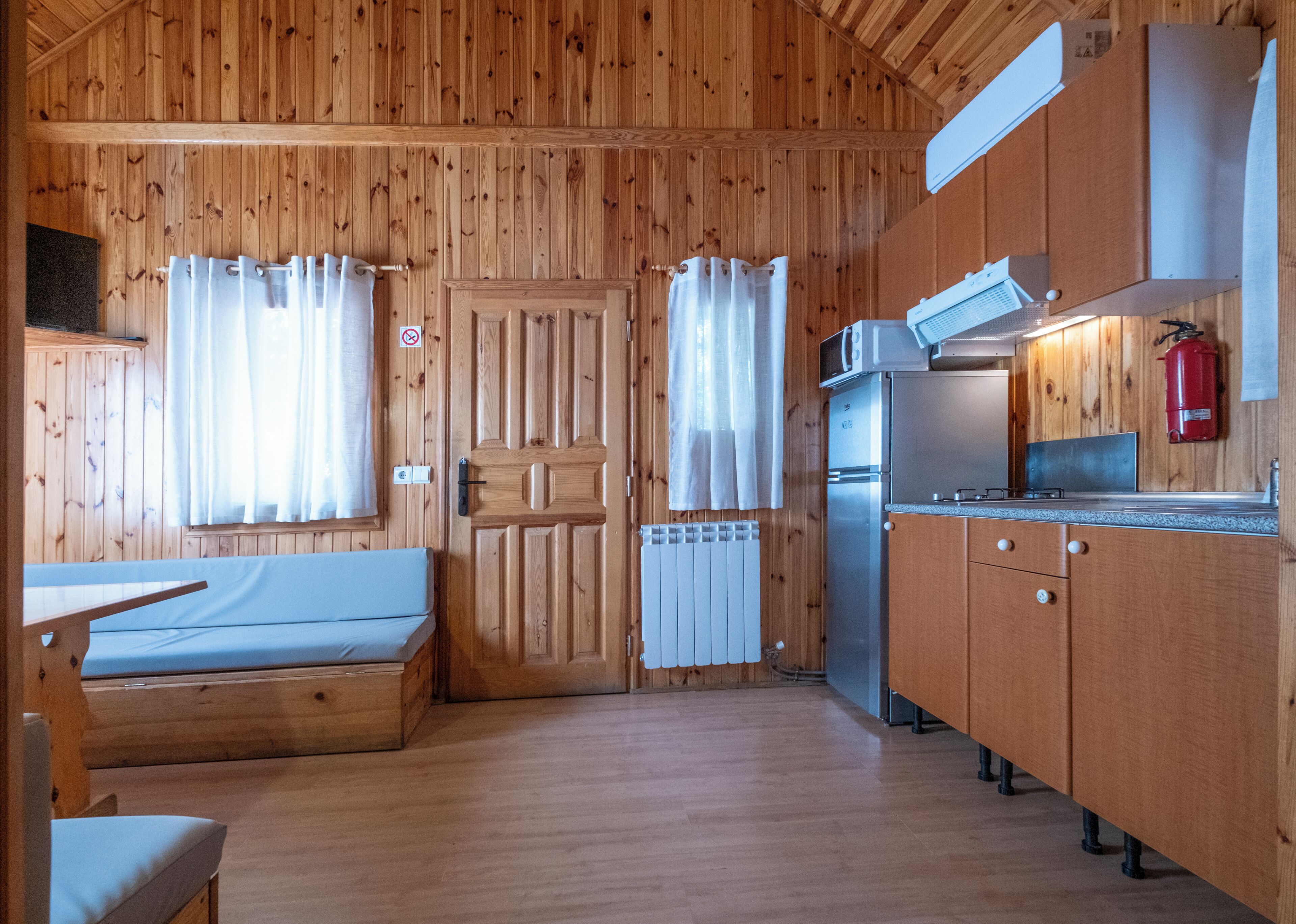 Interior de una cabaña de madera con kitchenette, una cama y una ventana con cortinas.