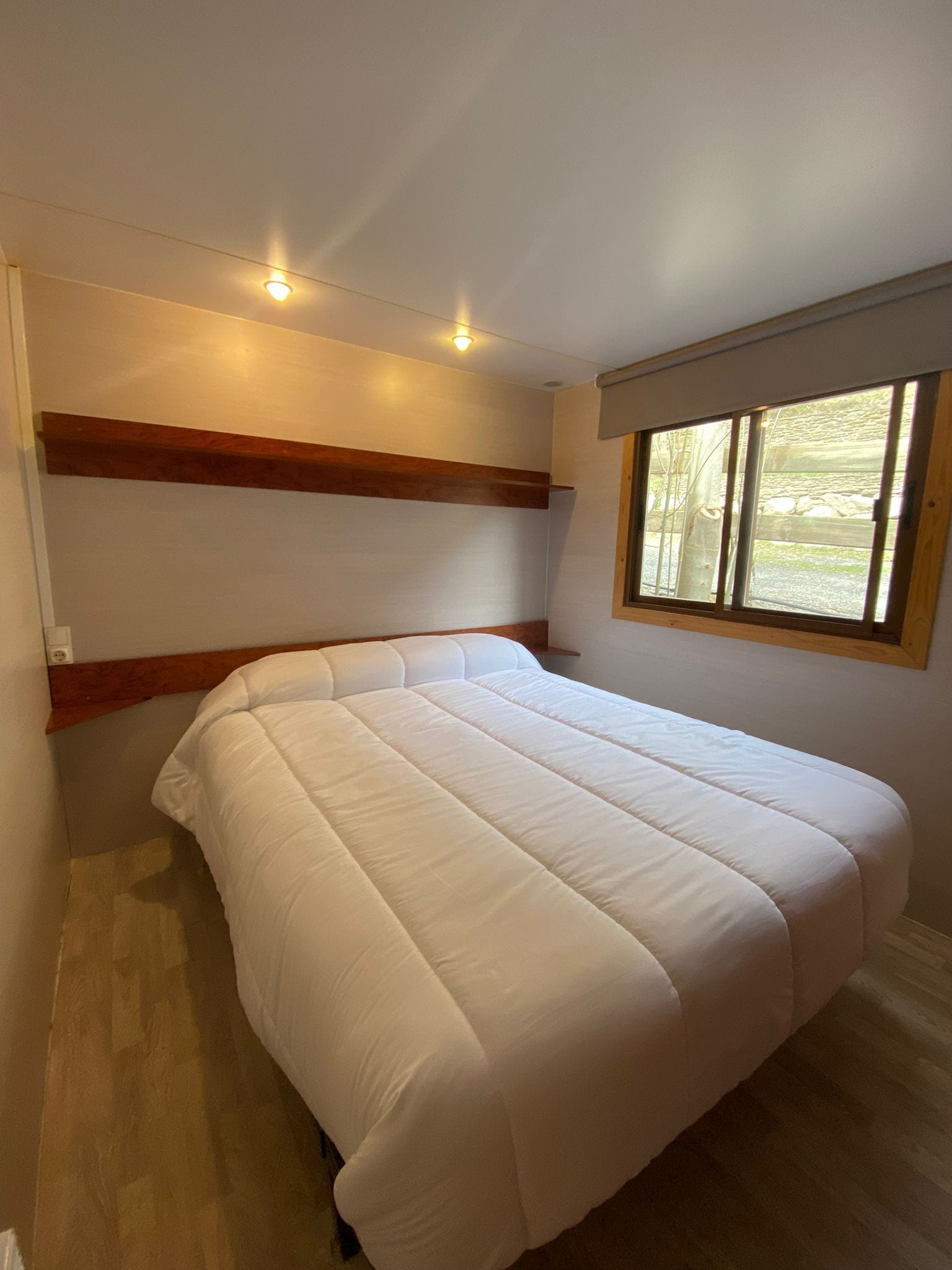 A bedroom with a bed, wooden headboard, a window with a view, and recessed ceiling lights.