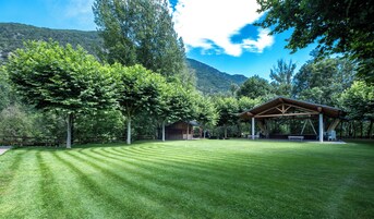 Un área verde bien cuidada con un pabellón de madera y una cabaña pequeña, rodeada de árboles y montañas.
