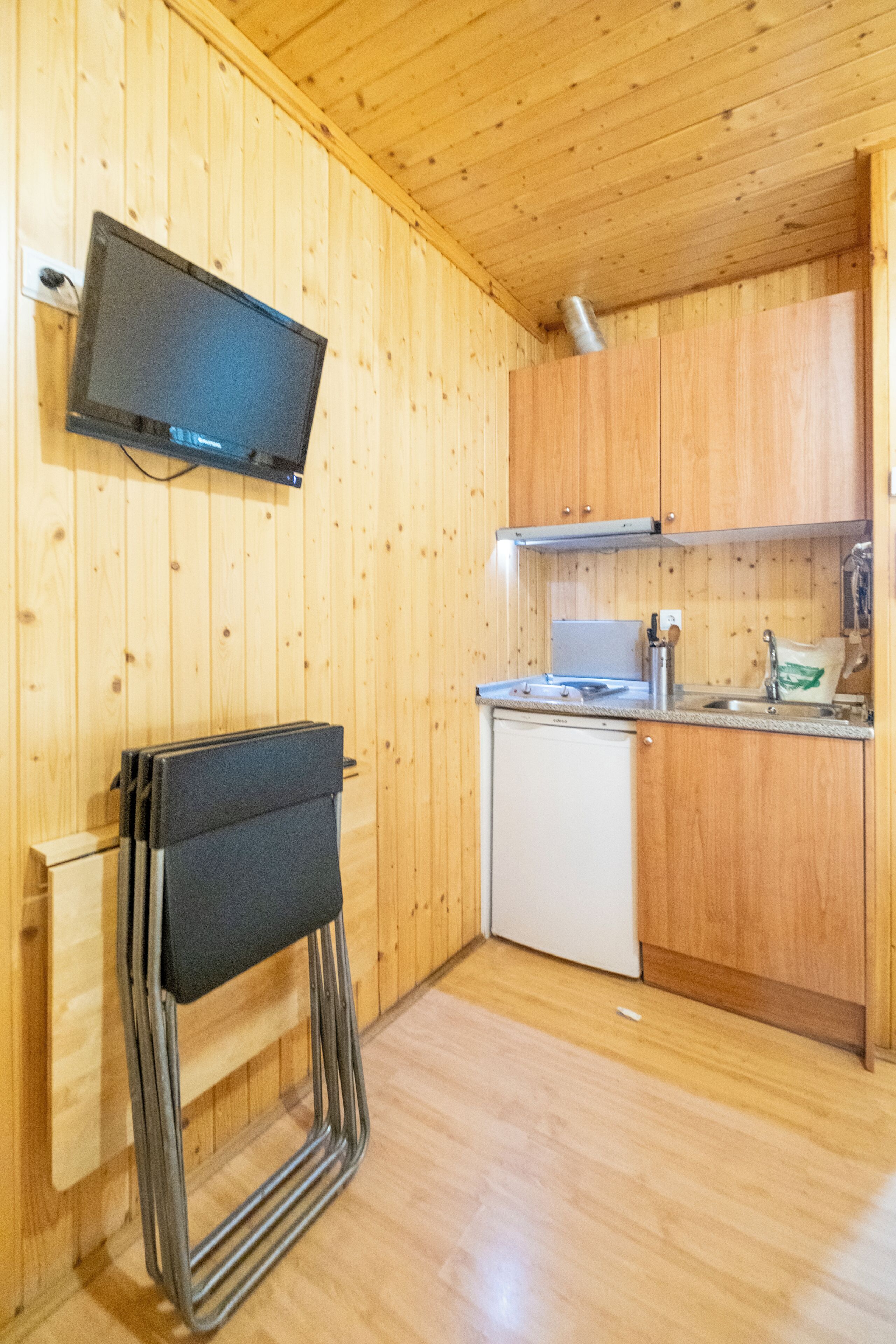 A compact kitchen with wooden walls, a built-in TV, a white dishwasher, and a metal chair.