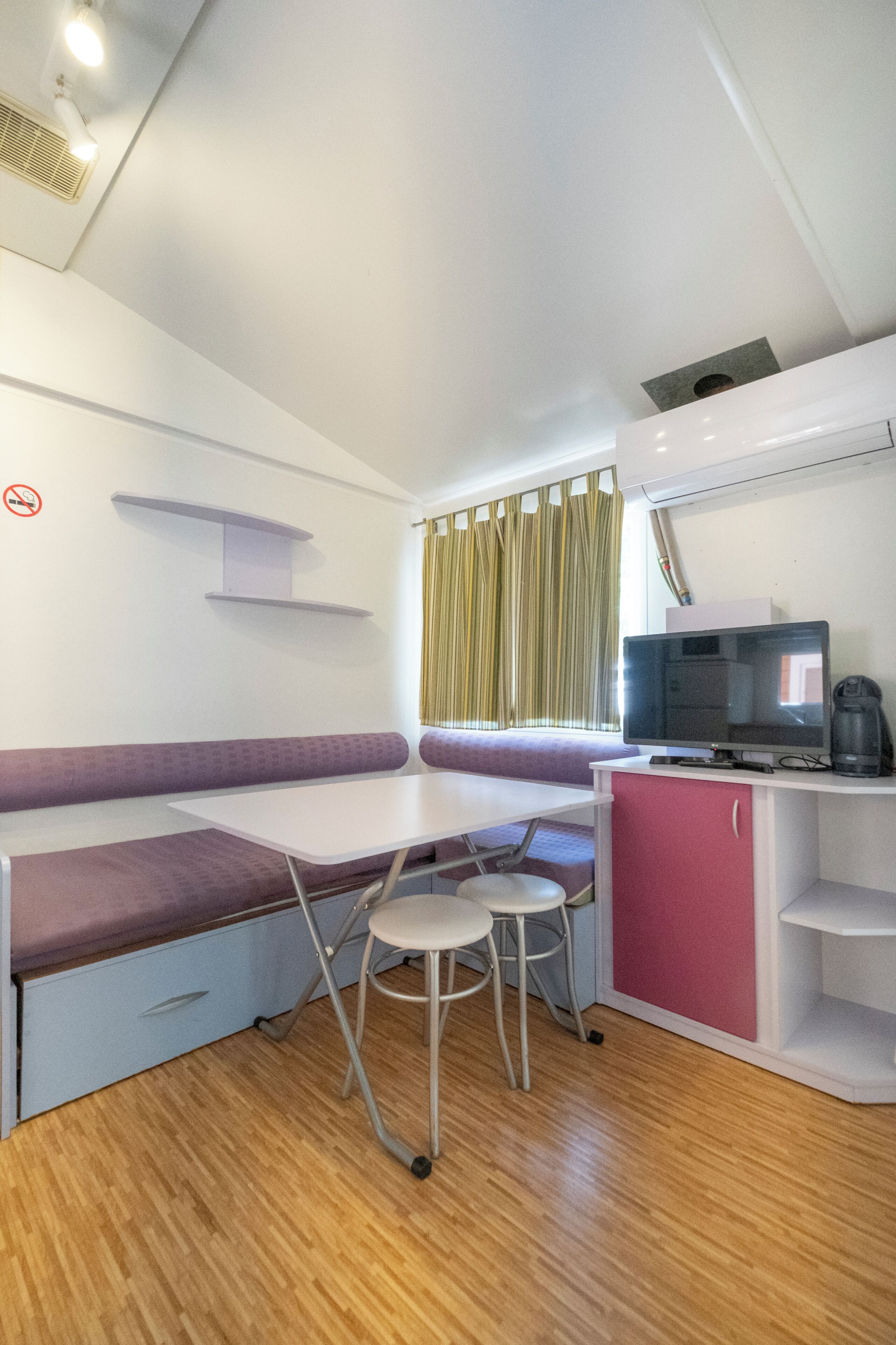 A compact room with a foldable table, a cushioned bench, a small stool, a television, and a cabinet.